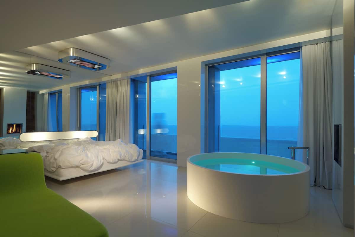 A modern hotel room with a large bed, a circular jacuzzi, and large windows offering a view of the sea.
