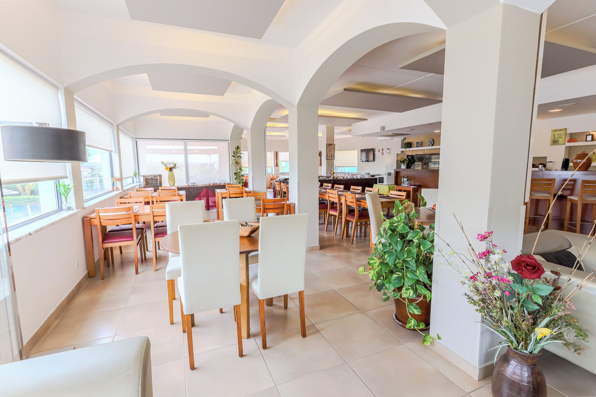 A spacious dining area with multiple tables and chairs, large windows, and indoor plants.