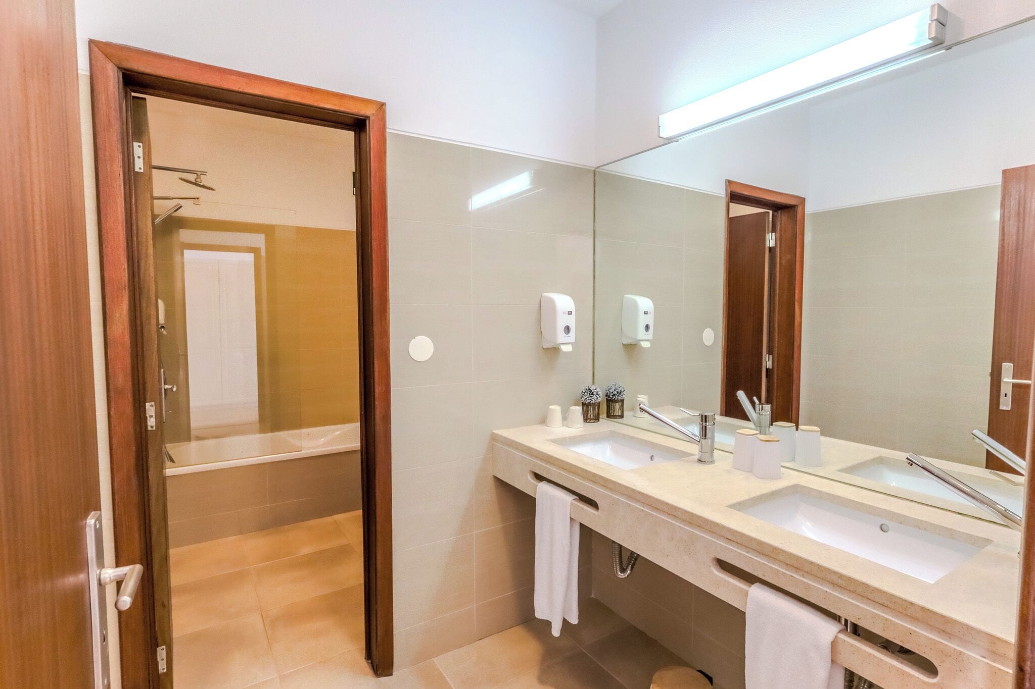 Un baño de hotel con lavamanos doble, un espejo grande, un perchero para toallas y una cabina de ducha con paredes de vidrio.