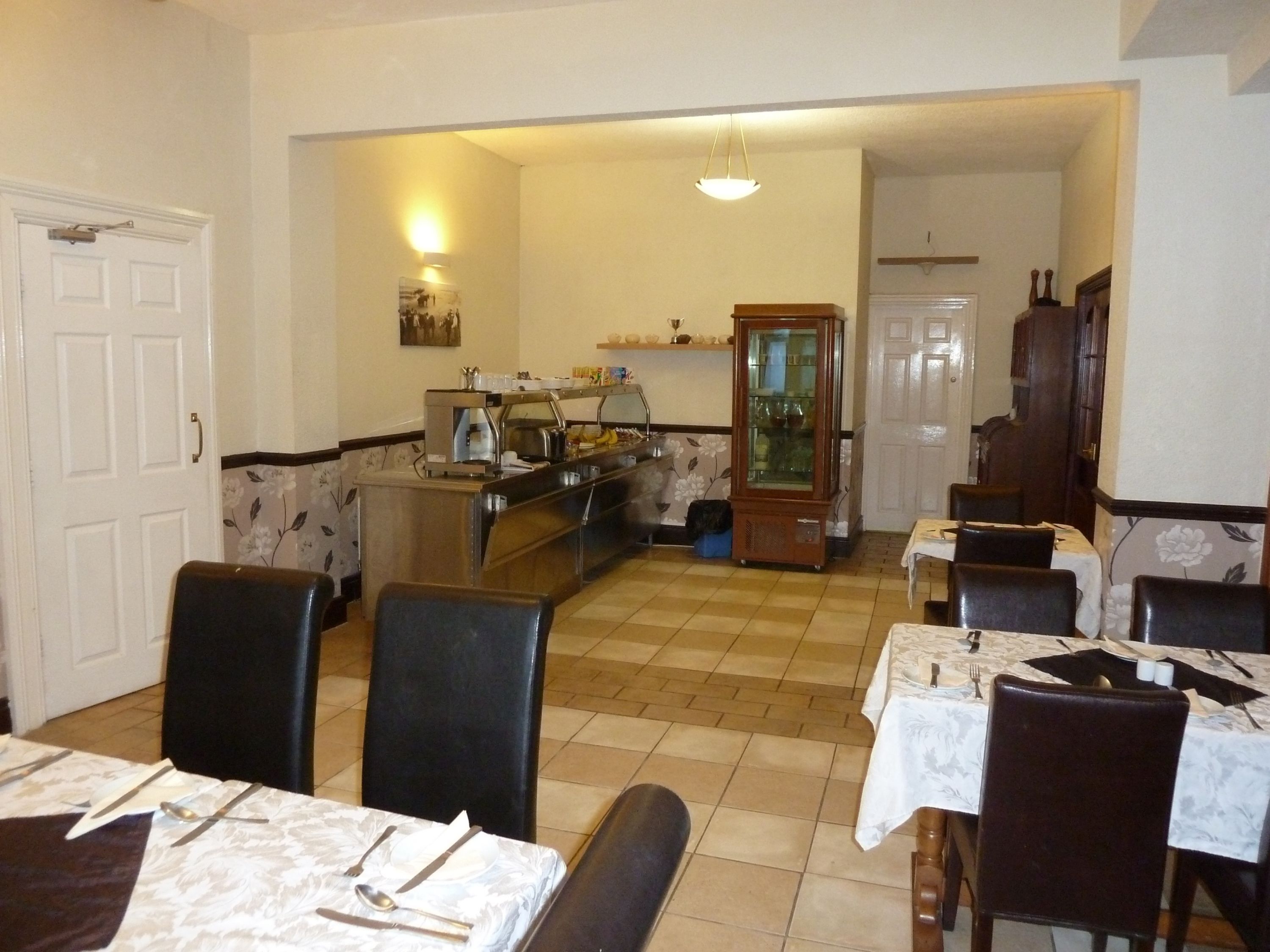 A dining area with tables set for meals, a kitchen area with a refrigerator and a coffee machine, and a cabinet with glassware.