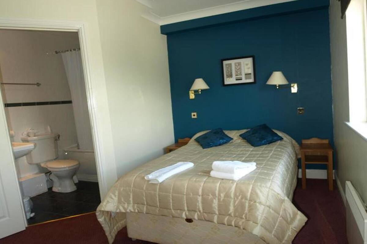 A bedroom with a bed, two bedside tables, a nightstand, a window, and a bathroom visible in the background.