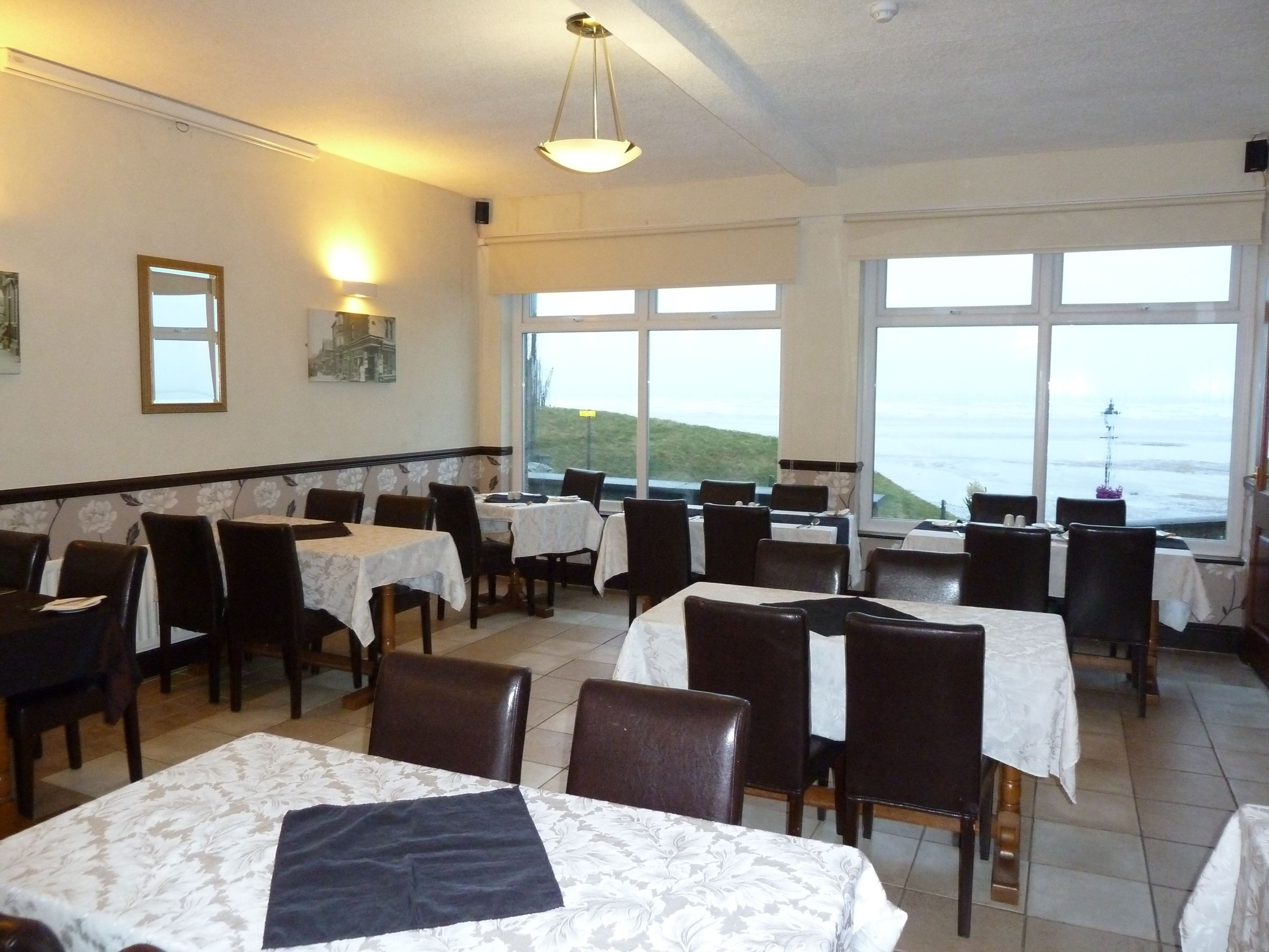 Un restaurant avec des tables et des chaises disposées pour accueillir les clients, de grandes fenêtres offrant une vue sur la mer et un luminaire au plafond.