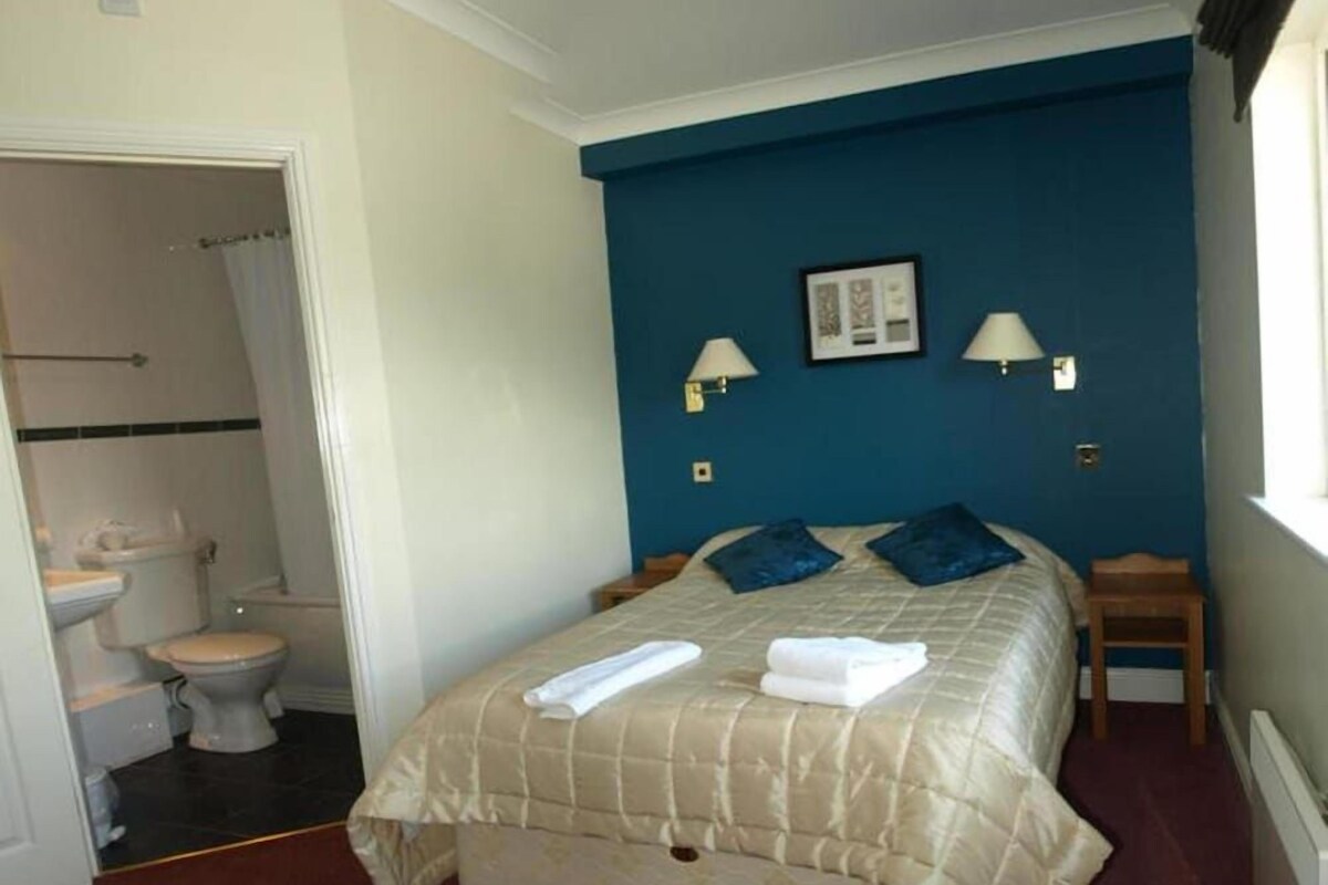A bedroom with a bed, two bedside tables, a window, a bathroom with a toilet and sink, and two wall-mounted lamps.