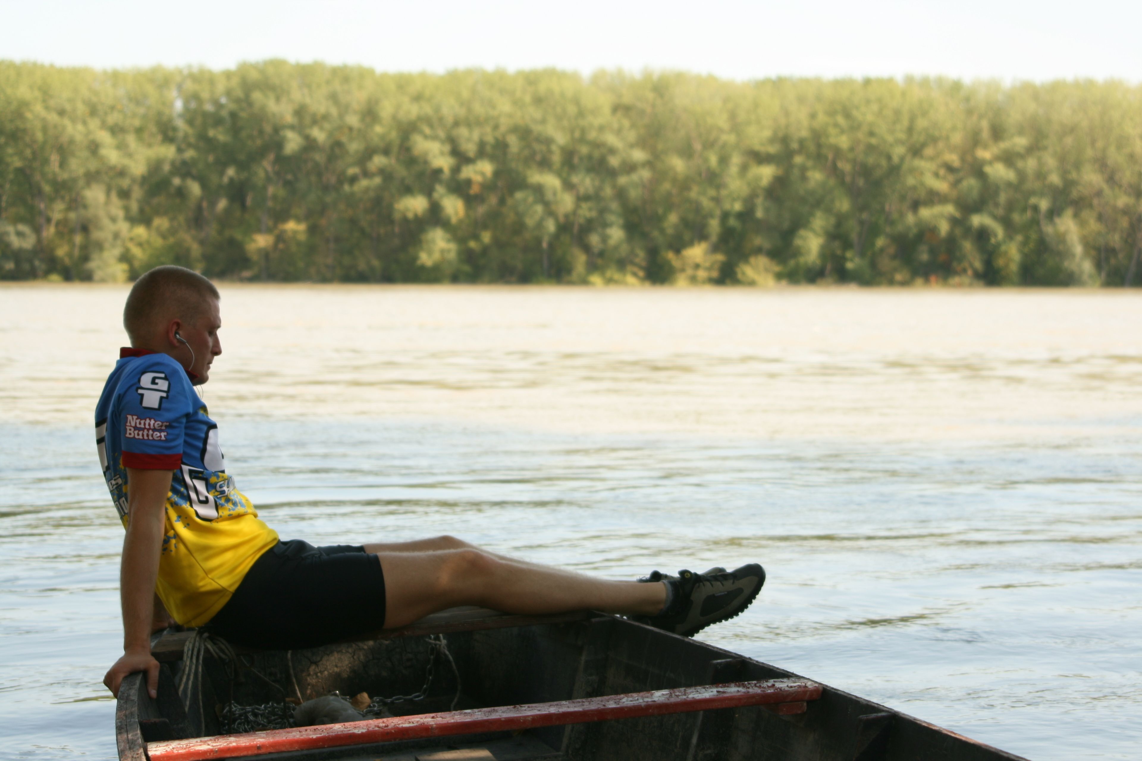 Une personne assise dans un bateau sur une étendue d’eau, avec une rive boisée en arrière-plan.