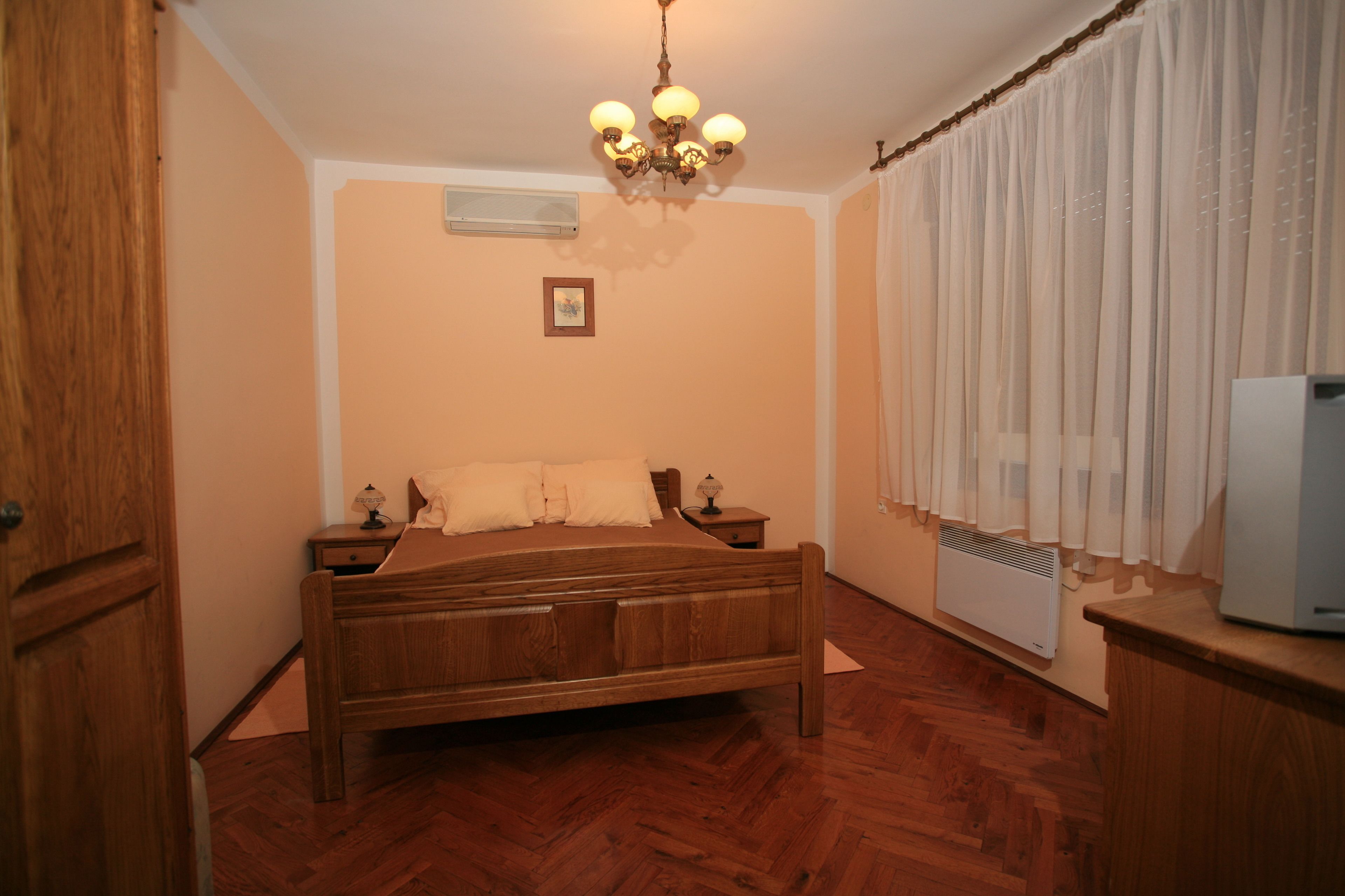 A bedroom with a wooden bed, a nightstand, a lamp, a ceiling light, a radiator, and a window with curtains.