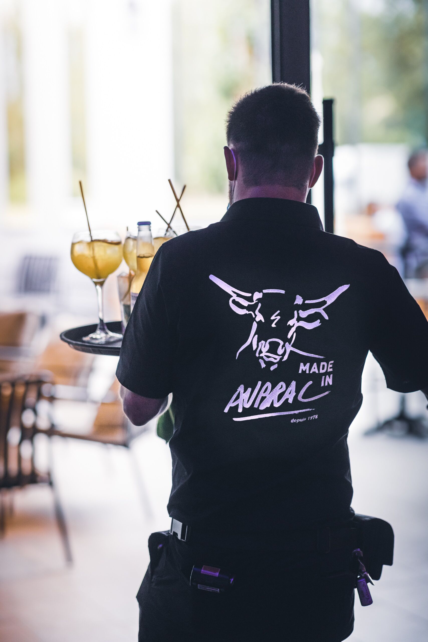 Une personne portant un t-shirt noir orné d’un motif graphique et d’un texte dans le dos, tenant un plateau rempli de boissons.