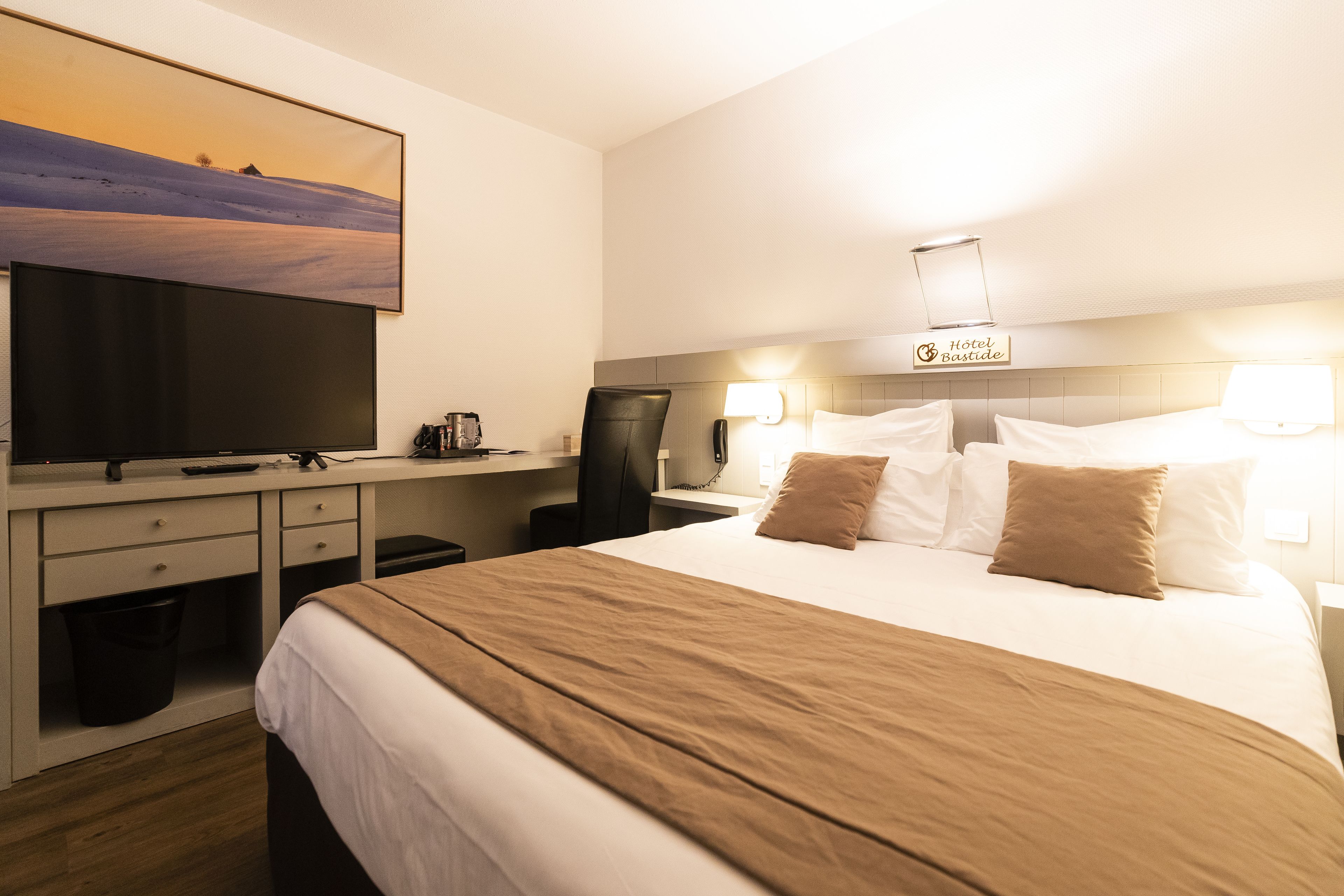 A hotel room with a bed, a desk with a computer, a TV, and a sign indicating the hotel name.