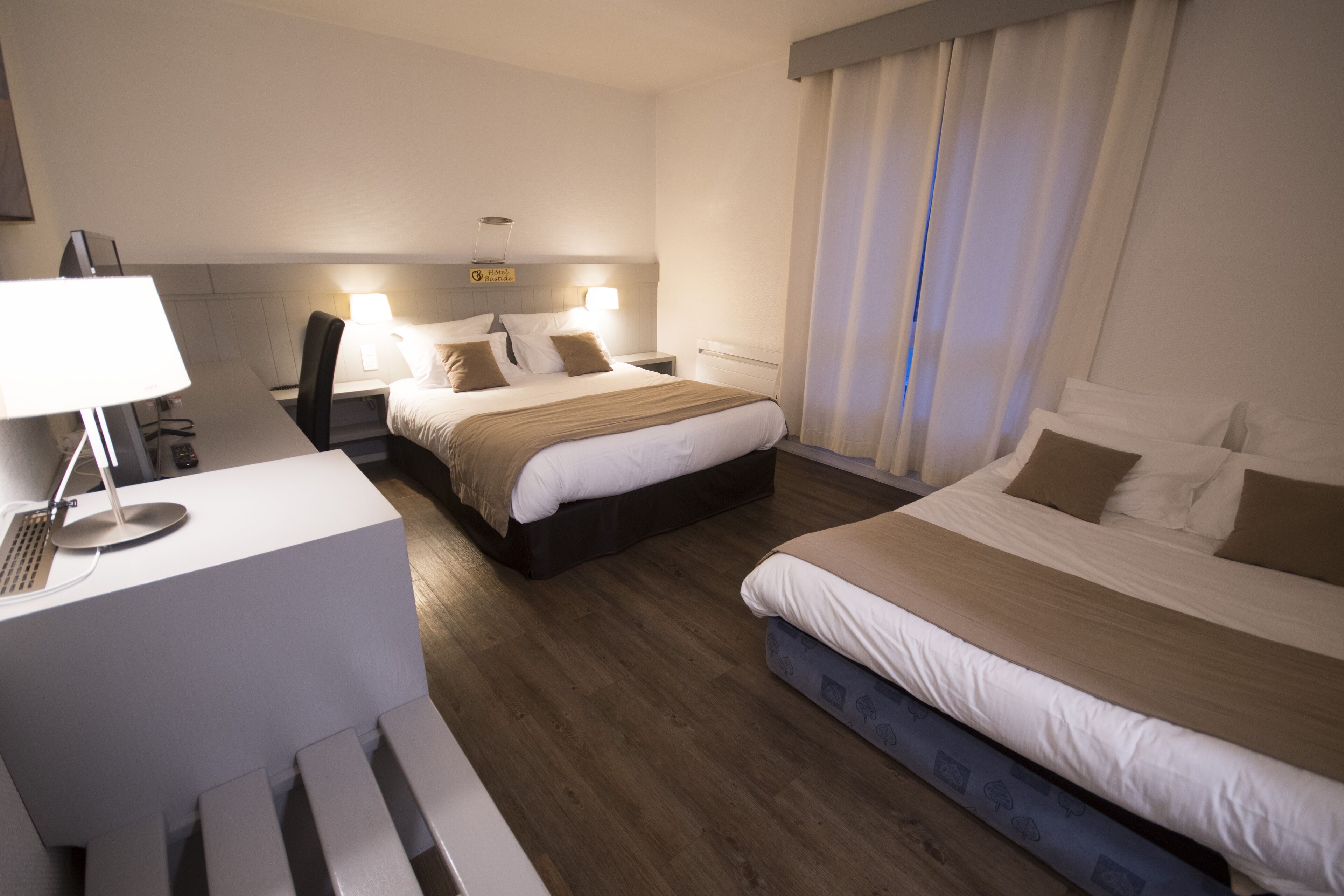 A hotel room with two beds, a desk with a lamp, a headboard with a sign, and a window with curtains.