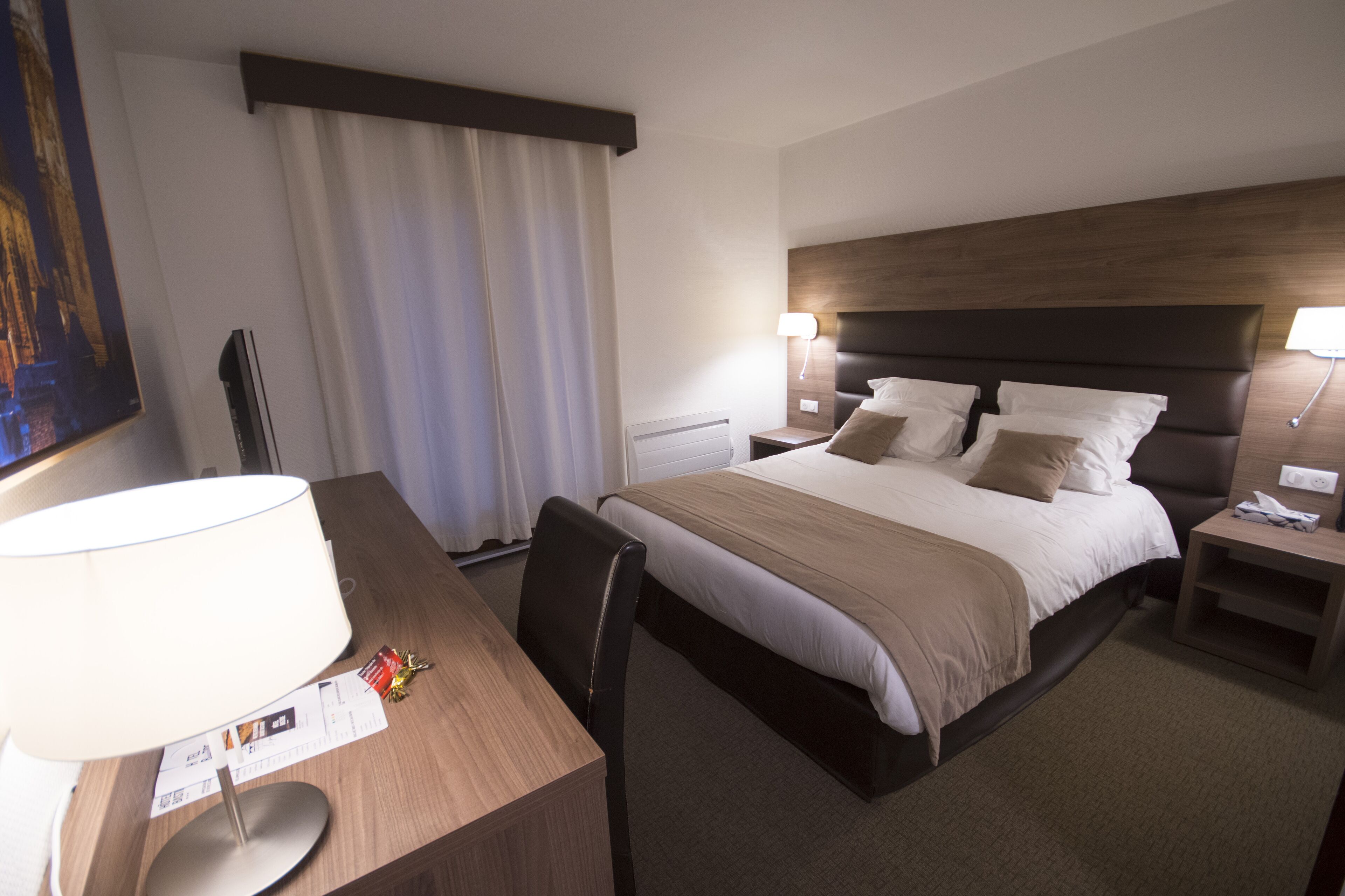 A hotel room with a bed, bedside tables, a desk, a chair, a lamp, and a painting on the wall.