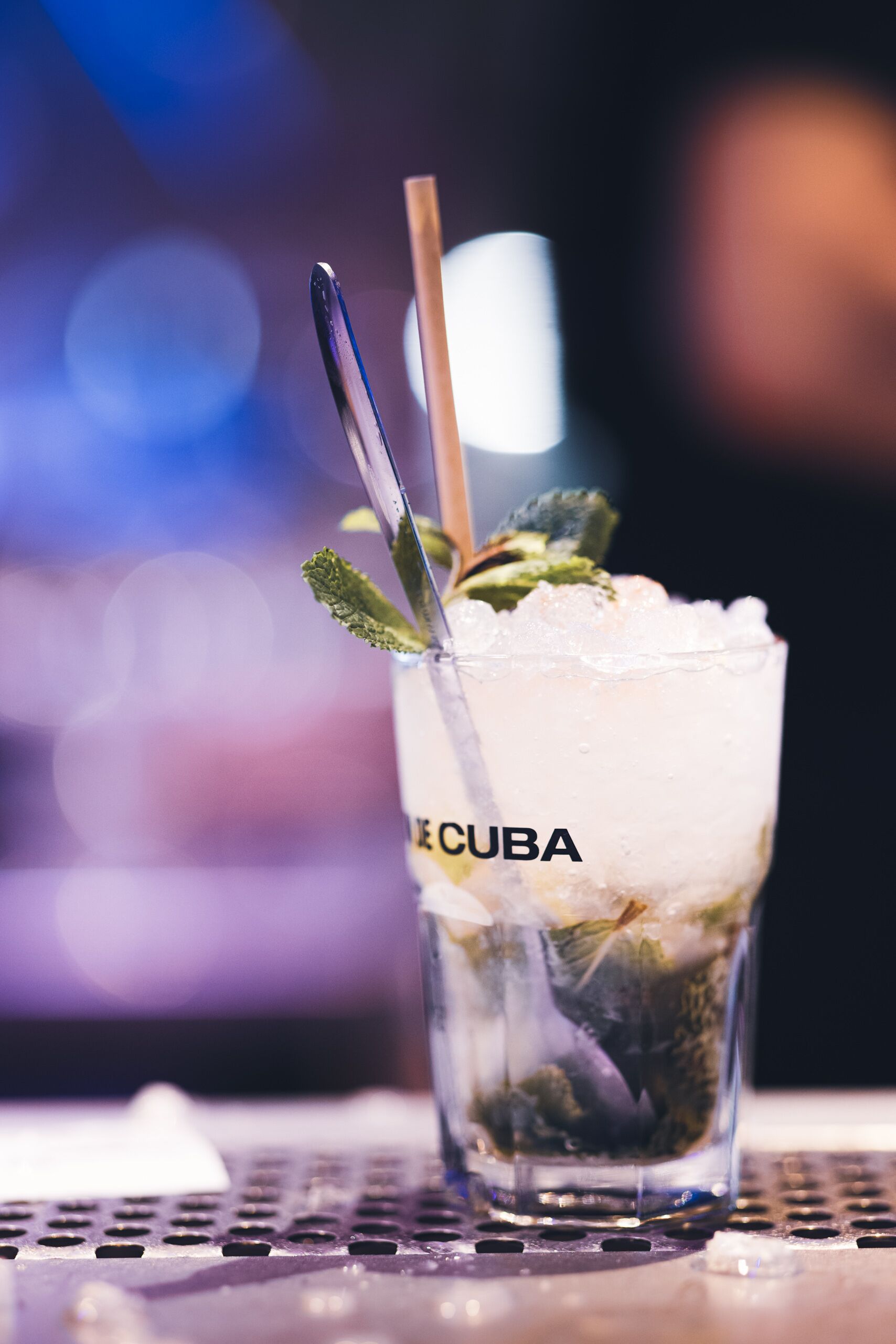 A glass of a beverage with a straw and mint garnish, labeled "CUBA," on a bar counter.