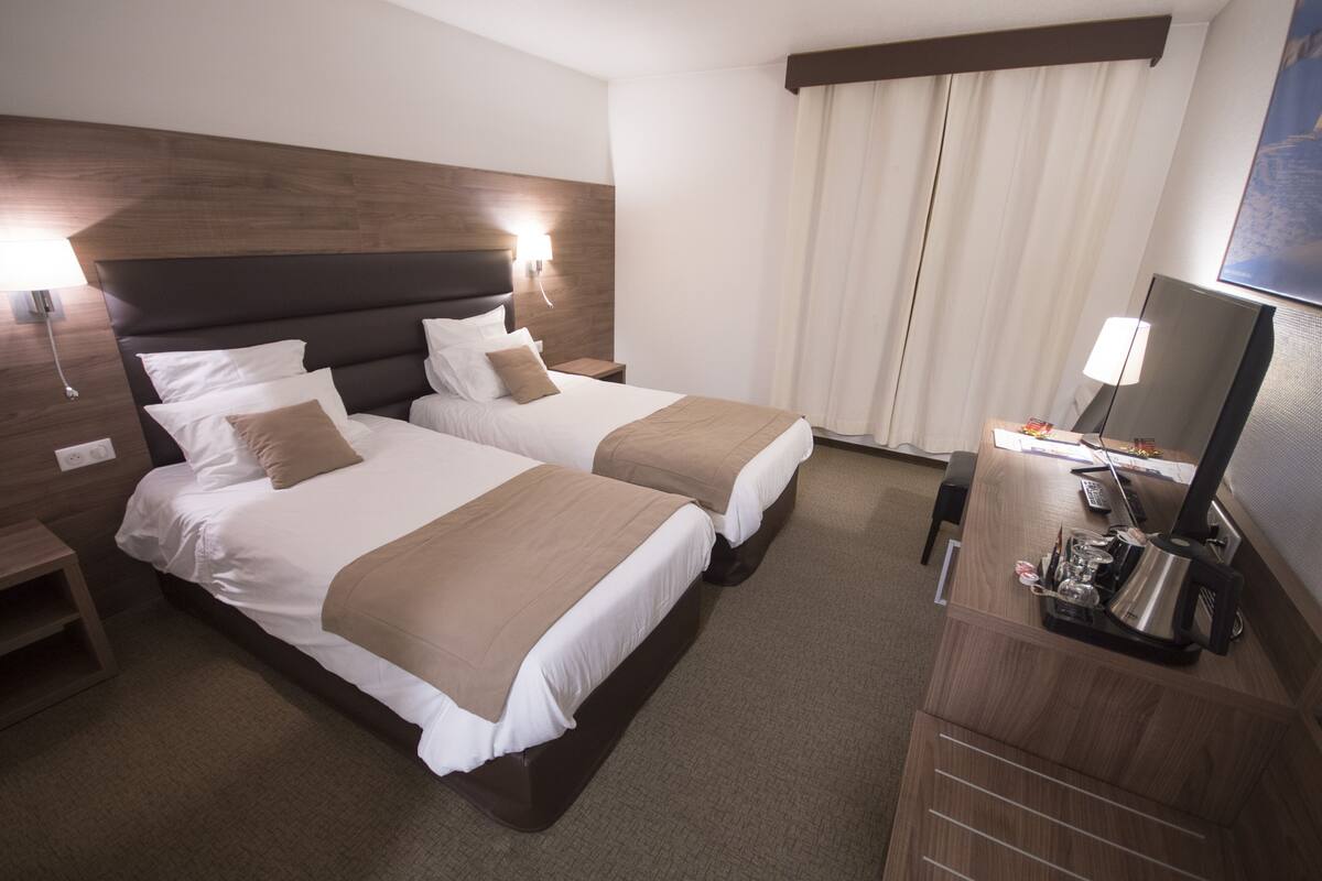 A hotel room with two beds, a desk with a computer, a lamp, and a window with curtains.