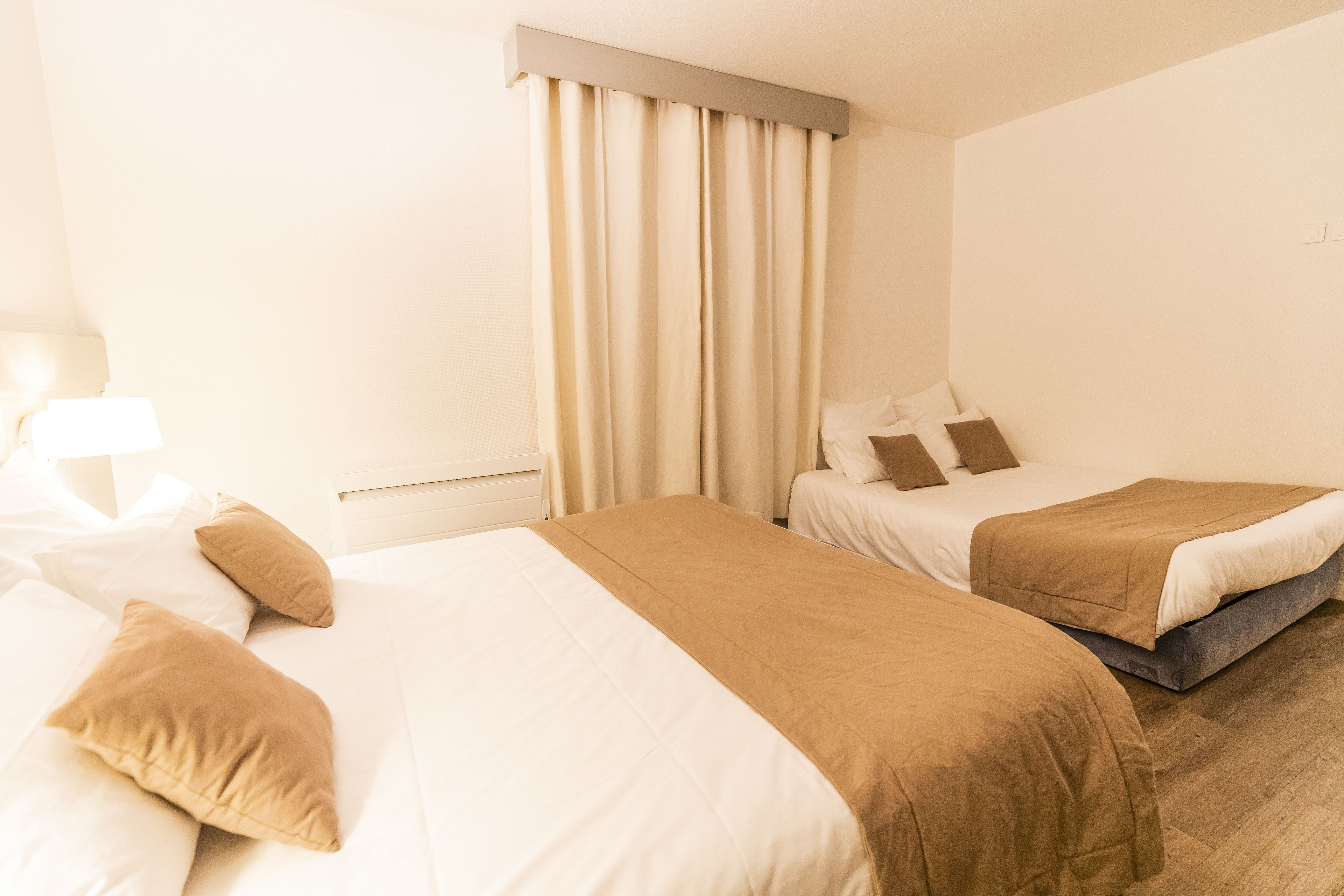 A hotel room with two beds, beige bedding, a window with curtains, and a wooden floor.