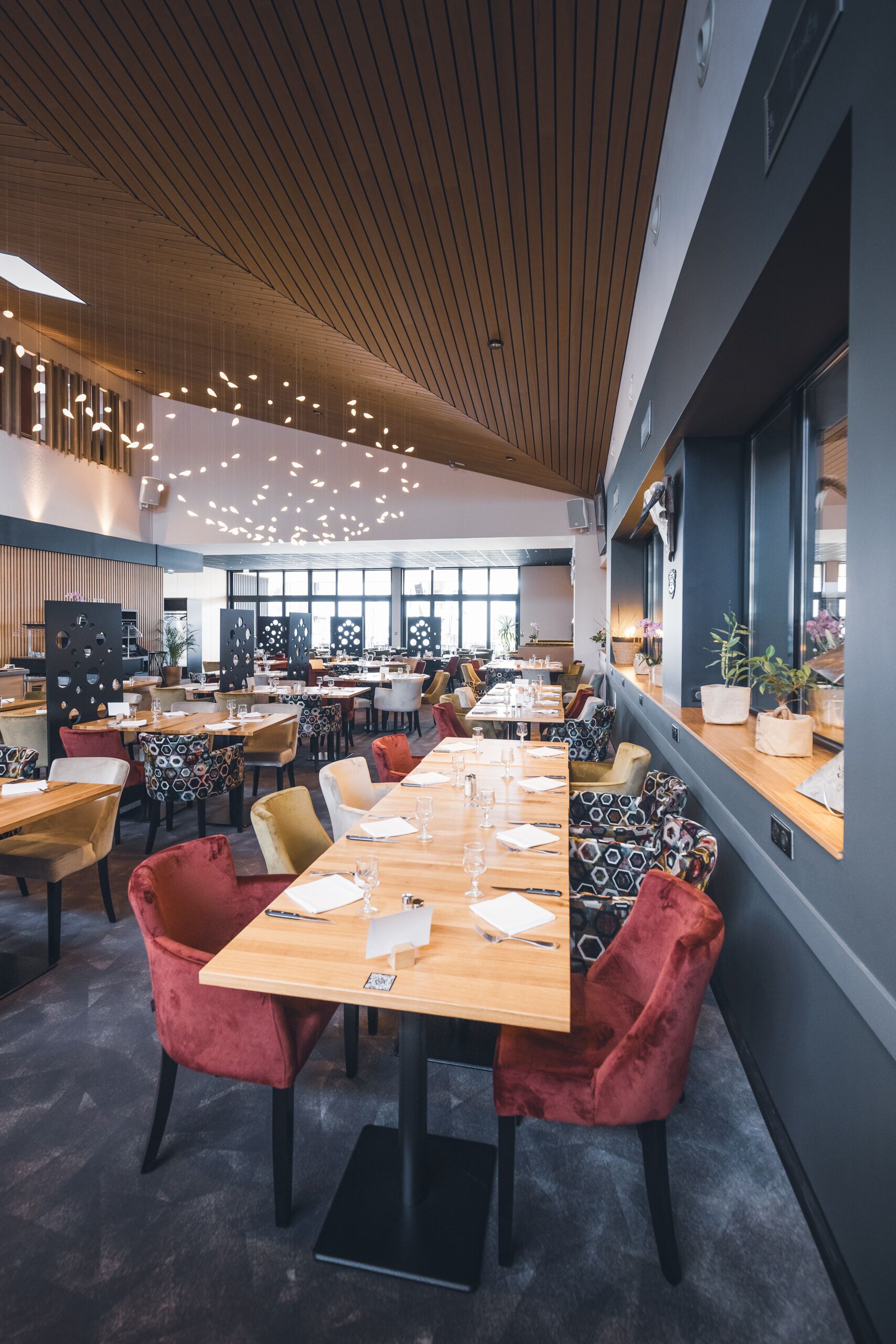 Un espace de restauration moderne avec de longues tables, des chaises en velours rouge et un plafond en bois.