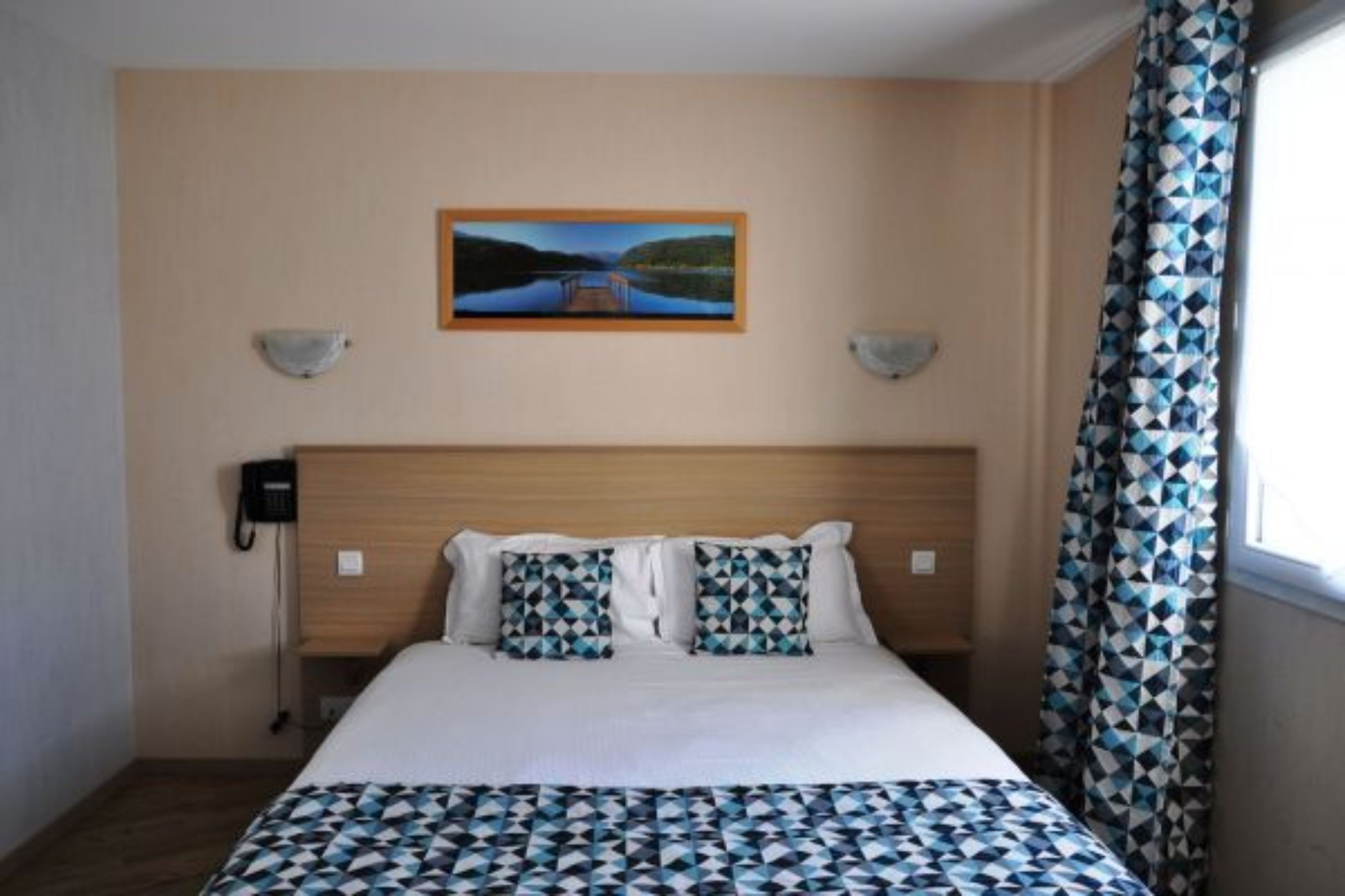 A hotel room with a bed, a painting on the wall, a telephone, and a patterned curtain.