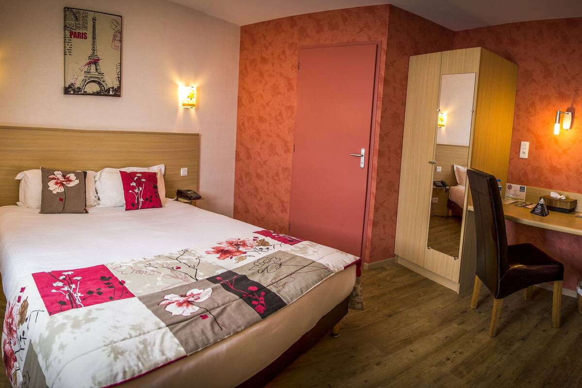 A hotel room with a bed, a desk, a chair, a mirror, and a television.