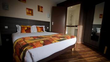 Une chambre d’hôtel avec un grand lit, un plancher en bois, une salle de bain visible à travers une porte ouverte et des œuvres d’art accrochées aux murs.