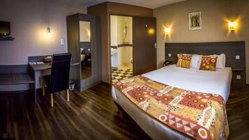 Habitación de hotel con cama, escritorio, silla, zona de ducha y televisor montado en la pared.