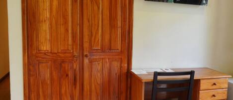 A room with a wooden wardrobe, a desk with a chair, a television mounted on the wall, and a trash bin.