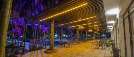Un patio al aire libre con mesas y sillas, rodeado de palmeras y una piscina.