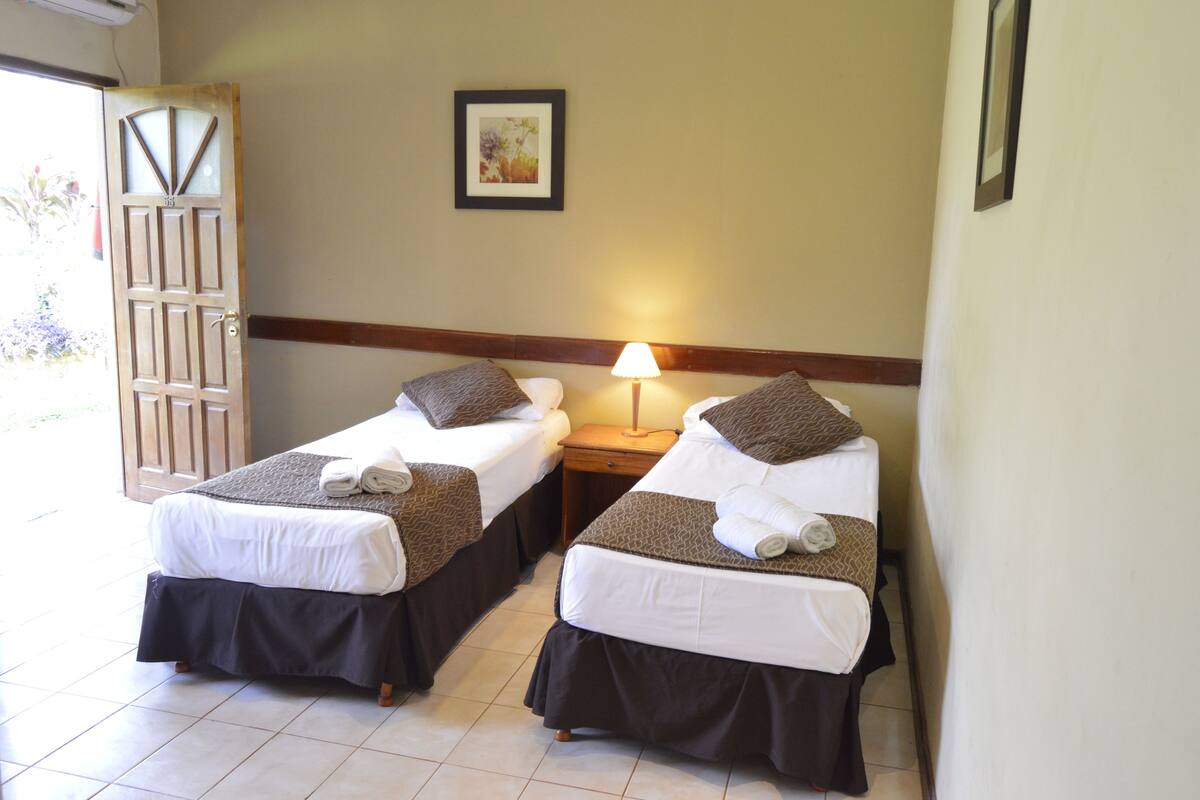 A hotel room with two single beds, each with a pillow and a blanket, a bedside table with a lamp, and a door leading outside.