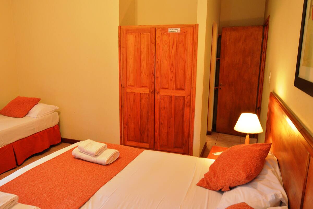 A hotel room with two beds, a wooden wardrobe, a nightstand with a lamp, and a red accent wall.