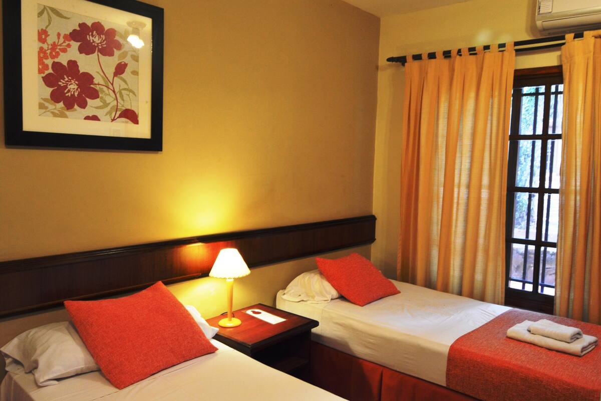 A hotel room with two beds, a nightstand with a lamp, a framed floral artwork on the wall, and a window with curtains.