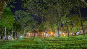 Un albergue con varias construcciones rodeado de árboles, que presenta una entrada bien iluminada y un área verde al frente.