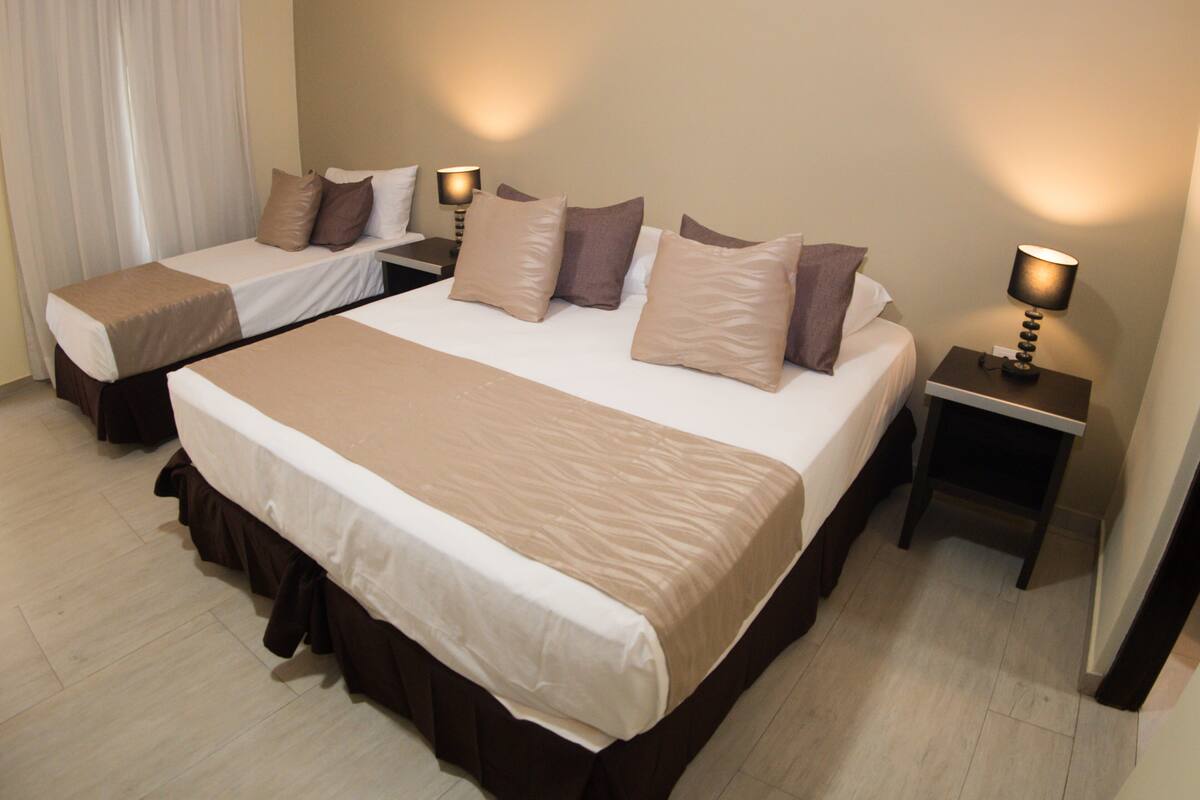 A hotel room with two beds, each with a brown bedspread and matching pillows. There are bedside tables with lamps and a window with curtains.