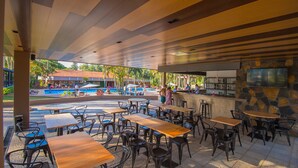 Un área de comedor al aire libre con mesas y sillas, una piscina al fondo y una zona de bar con pared de piedra y una pantalla de televisión.