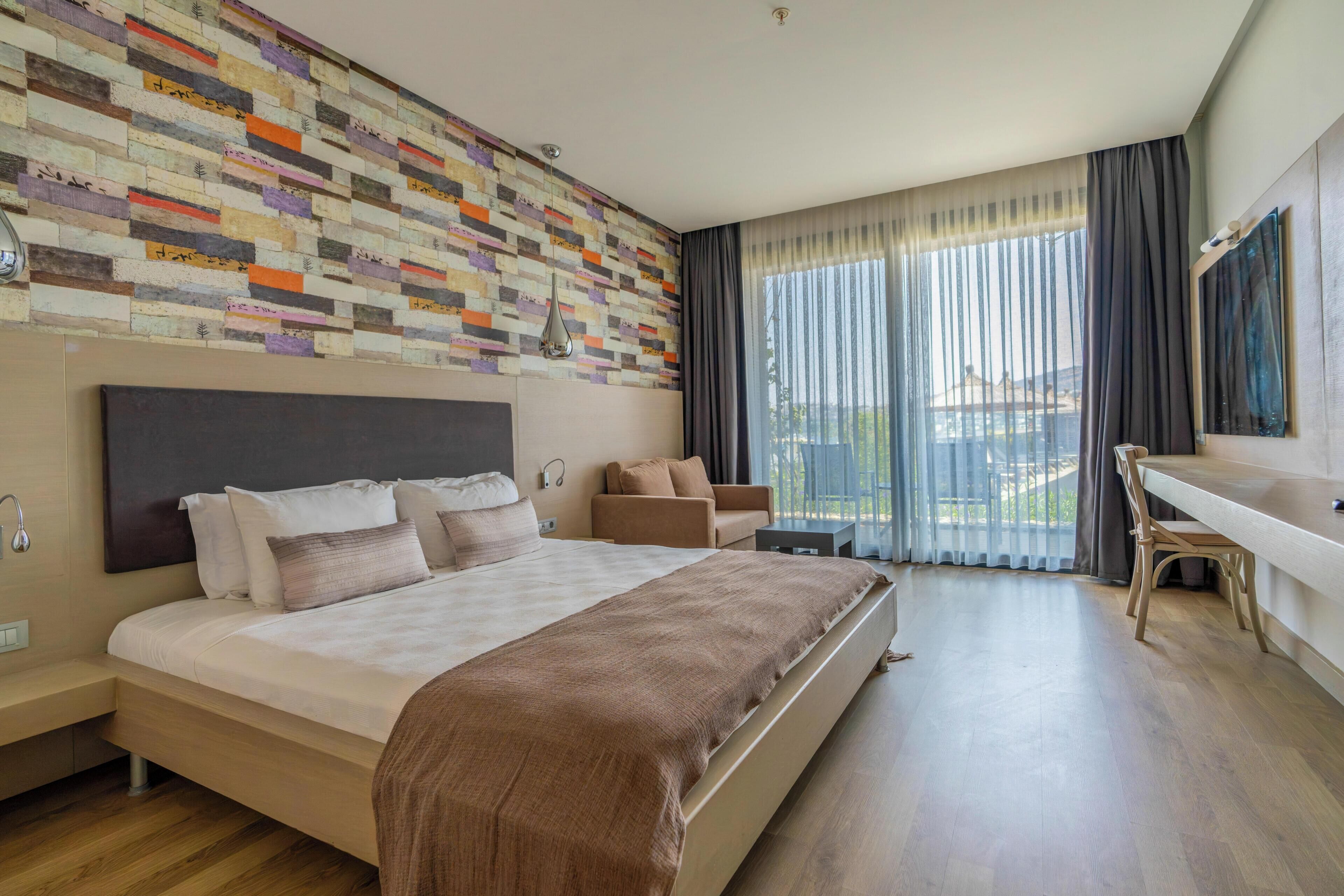 Habitación de hotel moderna con una cama grande, un escritorio y vistas de la ciudad a través de cortinas translúcidas.