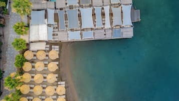 Vista aérea de un complejo turístico frente a la playa, con múltiples tumbonas y sombrillas, una terraza de madera y una vista despejada del océano.