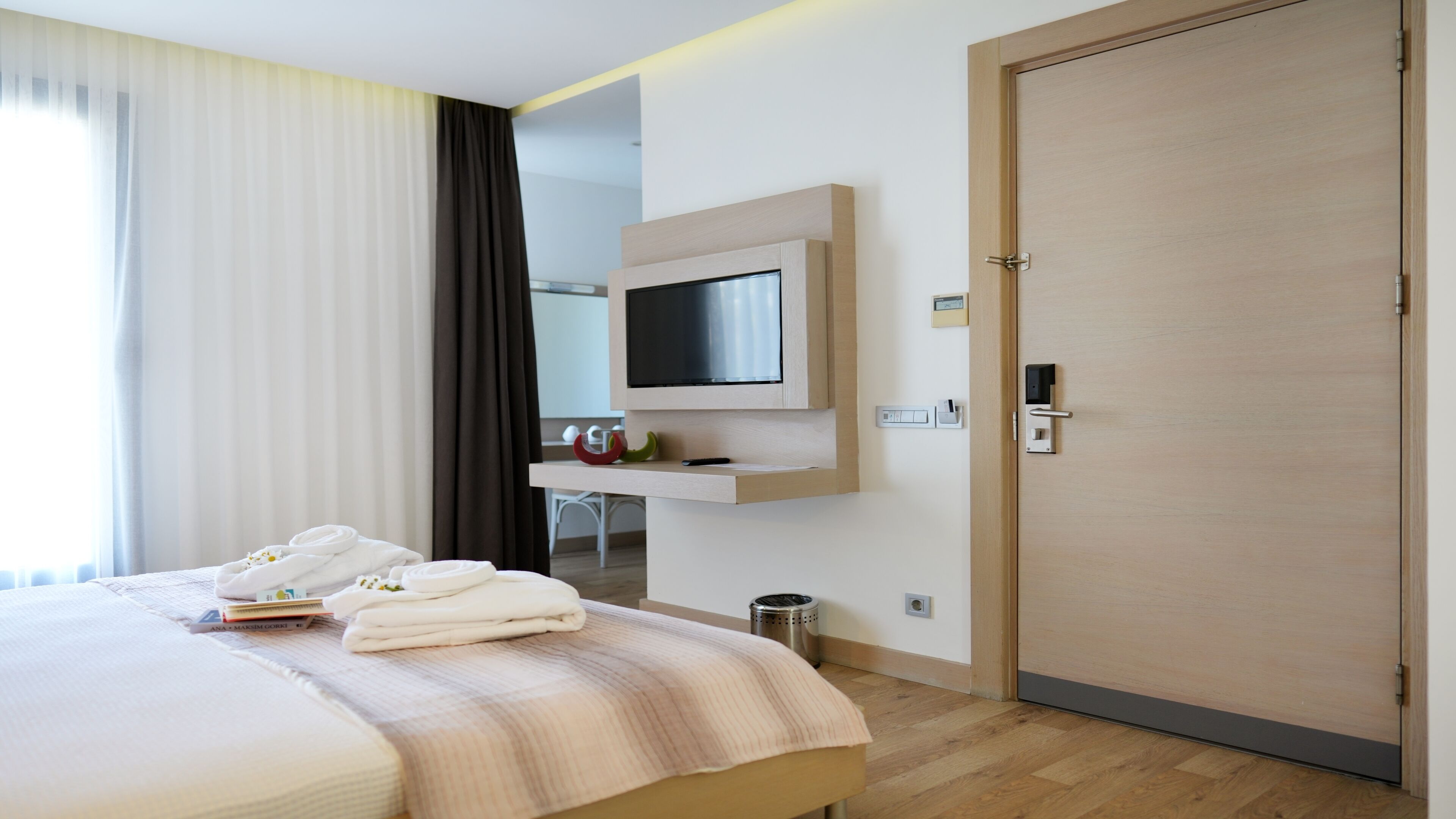 A hotel room with a bed, a TV mounted on the wall, a door with a handle, and a small trash bin.