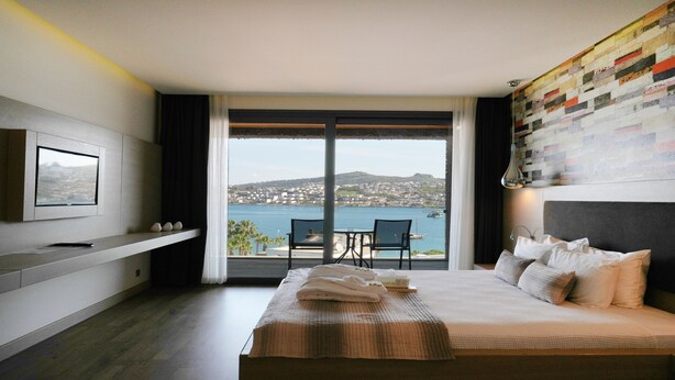 A modern hotel room with a large bed, a TV mounted on the wall, a balcony with a view of the sea and a cityscape. - Costa Farilya Special Class Hotel Bodrum - Special Class (Bodrum)