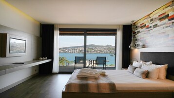 A modern hotel room with a large bed, a TV mounted on the wall, a balcony with a view of the sea and a cityscape.