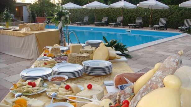 Montaje de buffet con diversos quesos, frutas y platos, dispuesto sobre una mesa cerca de una zona de piscina con sillones y sombrillas.