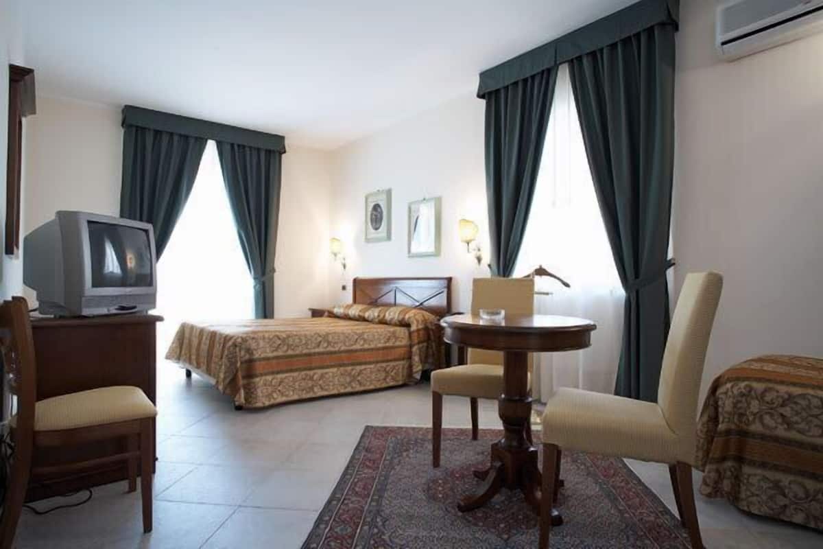 Une chambre d’hôtel avec un lit, une chaise, une petite table, un téléviseur et deux fenêtres avec des rideaux.