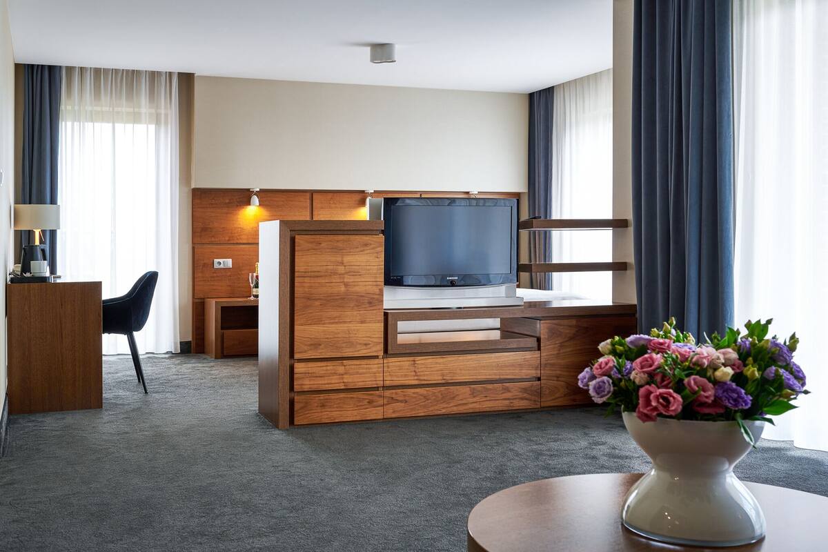 A hotel room with a wooden desk, a flat-screen TV, a vase of flowers, and a round table.