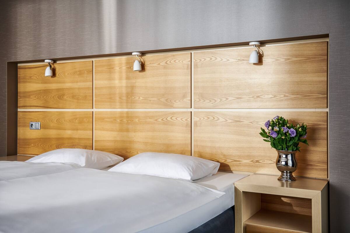 A hotel room with a wooden headboard, two white beds, a bedside table with a vase of flowers, and a wall-mounted light fixture.