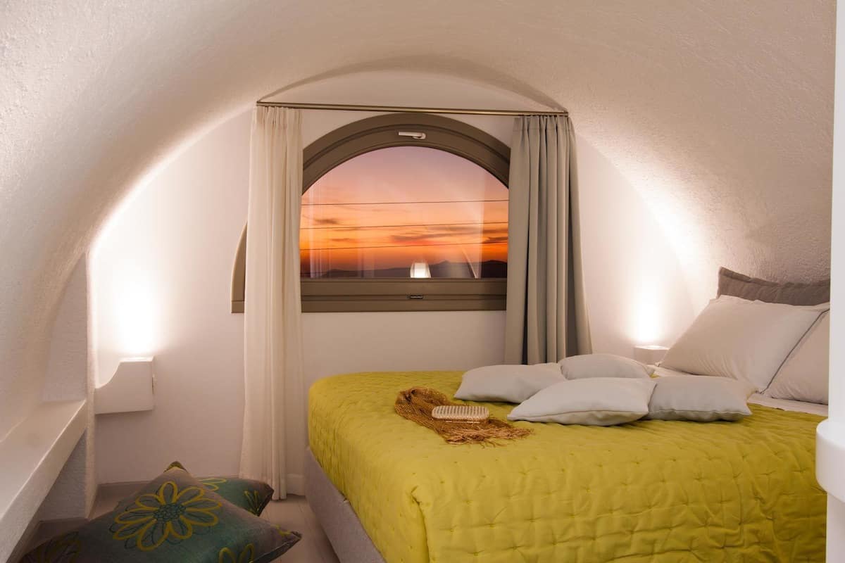 Une chambre à coucher avec un lit, des oreillers, une fenêtre avec des rideaux et une vue sur un coucher de soleil.