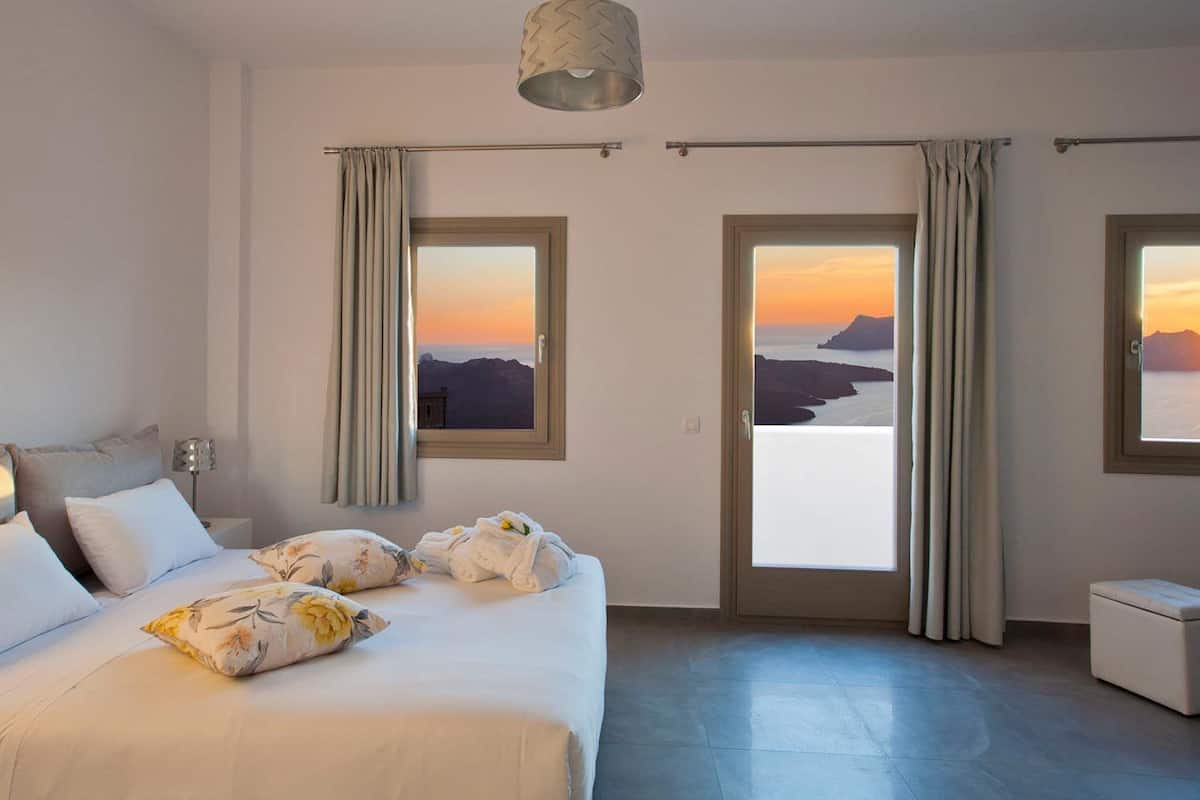 Une chambre d’hôtel avec un grand lit, deux fenêtres avec des rideaux, une vue sur le coucher du soleil et une petite table de chevet.
