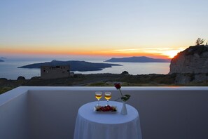 Una mesa con mantel blanco, dos copas de vino, una rosa y un plato de fruta en un balcón con vistas a un atardecer y un paisaje costero.
