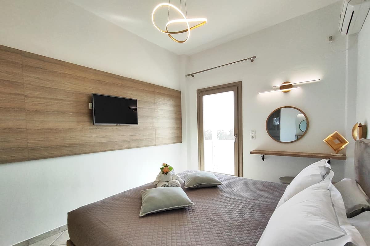 Une chambre moderne avec un grand lit, un téléviseur fixé au mur, un mur recouvert de panneaux en bois, une porte donnant sur l’extérieur et une petite étagère avec un miroir et un cadre photo.