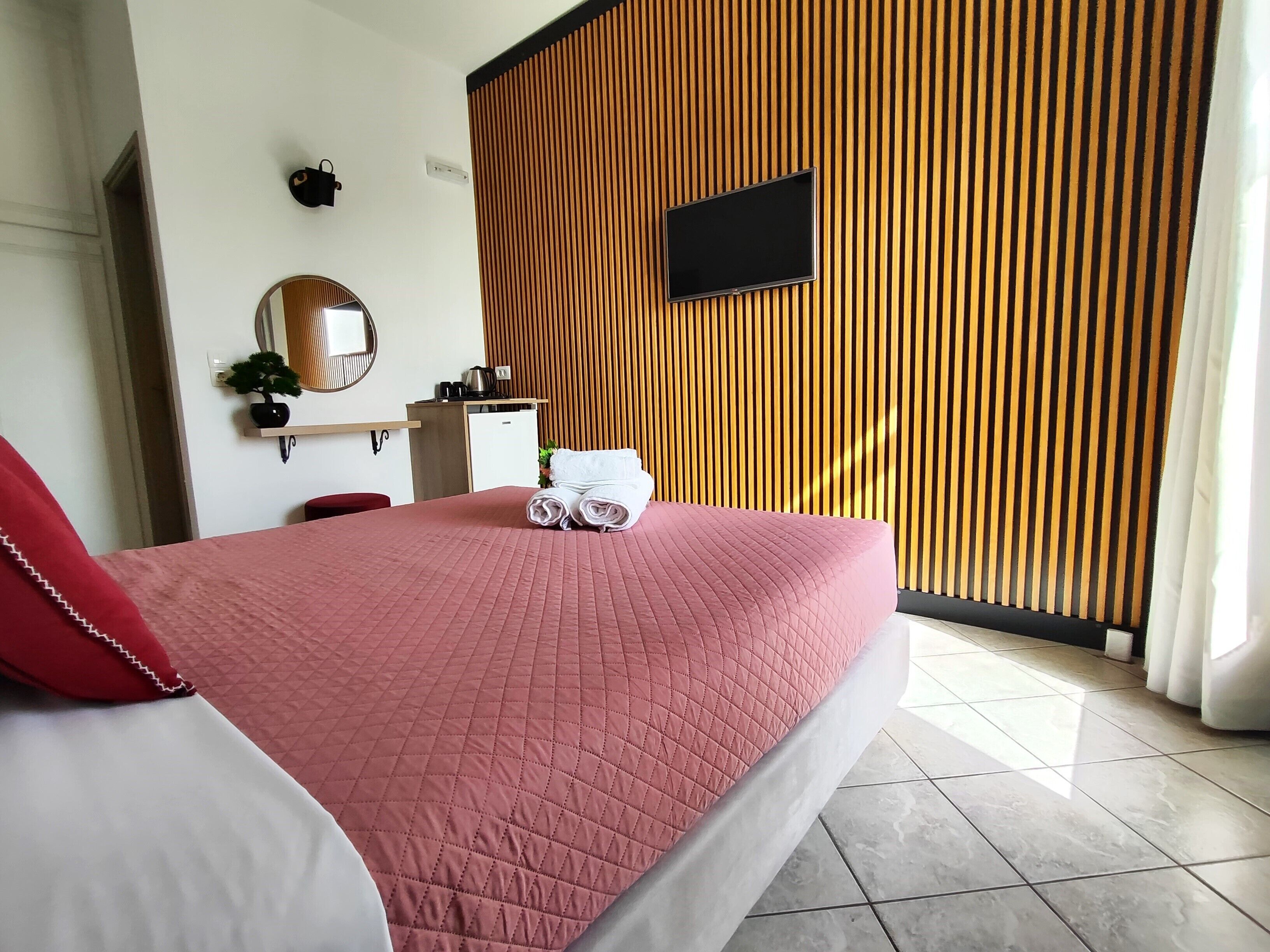 Una habitació d'hotel amb un llit, una televisió penjada a un panell de paret de fusta, un escriptori petit amb un bullidor i una cadira vermella.