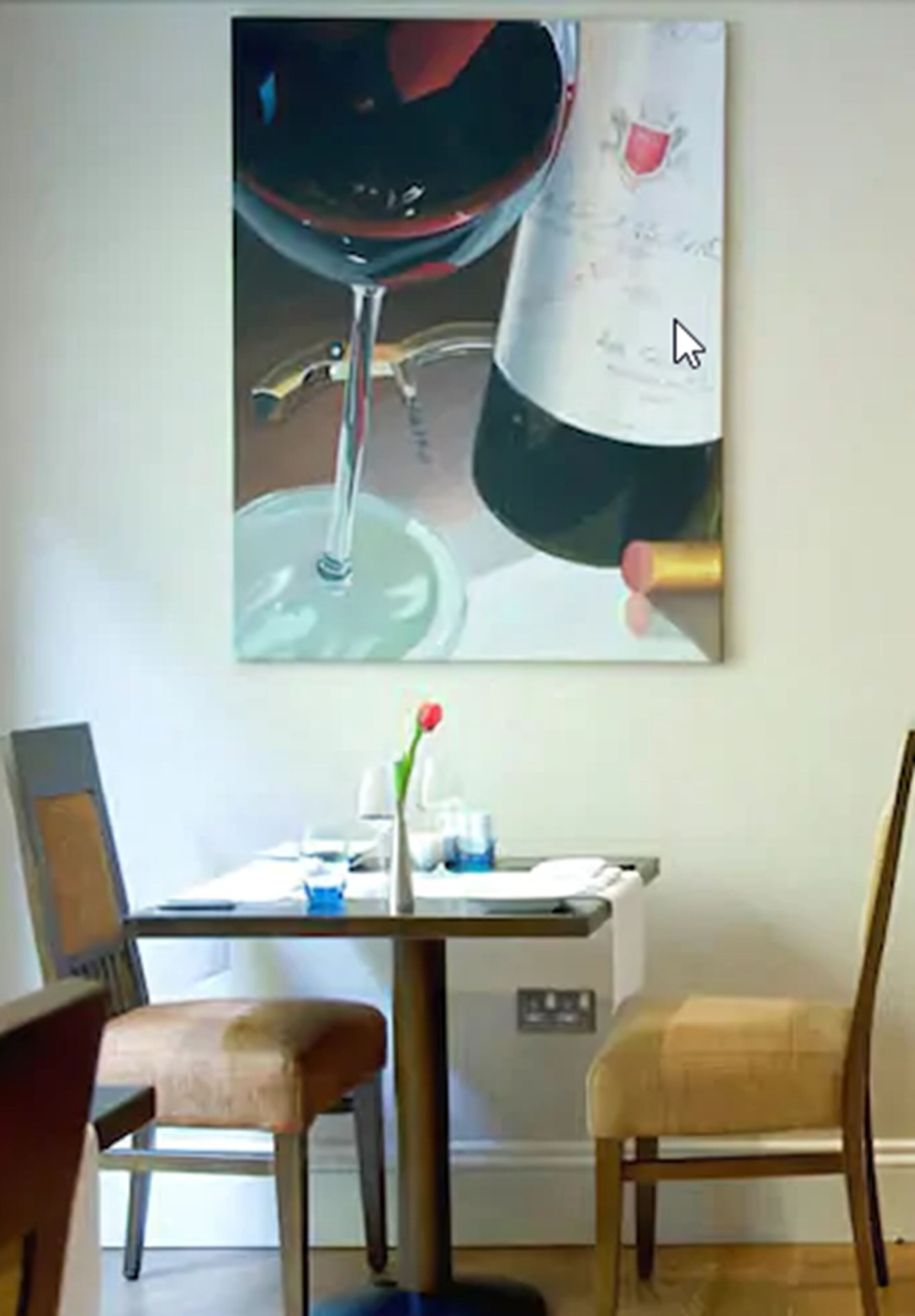 A dining area with a table, chairs, a vase with a red flower, and a painting of a wine glass on the wall.