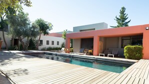 A modern outdoor pool area with a wooden deck, lounge chairs, and a building with large windows. - Marianne Wine Estate (Stellenbosch)