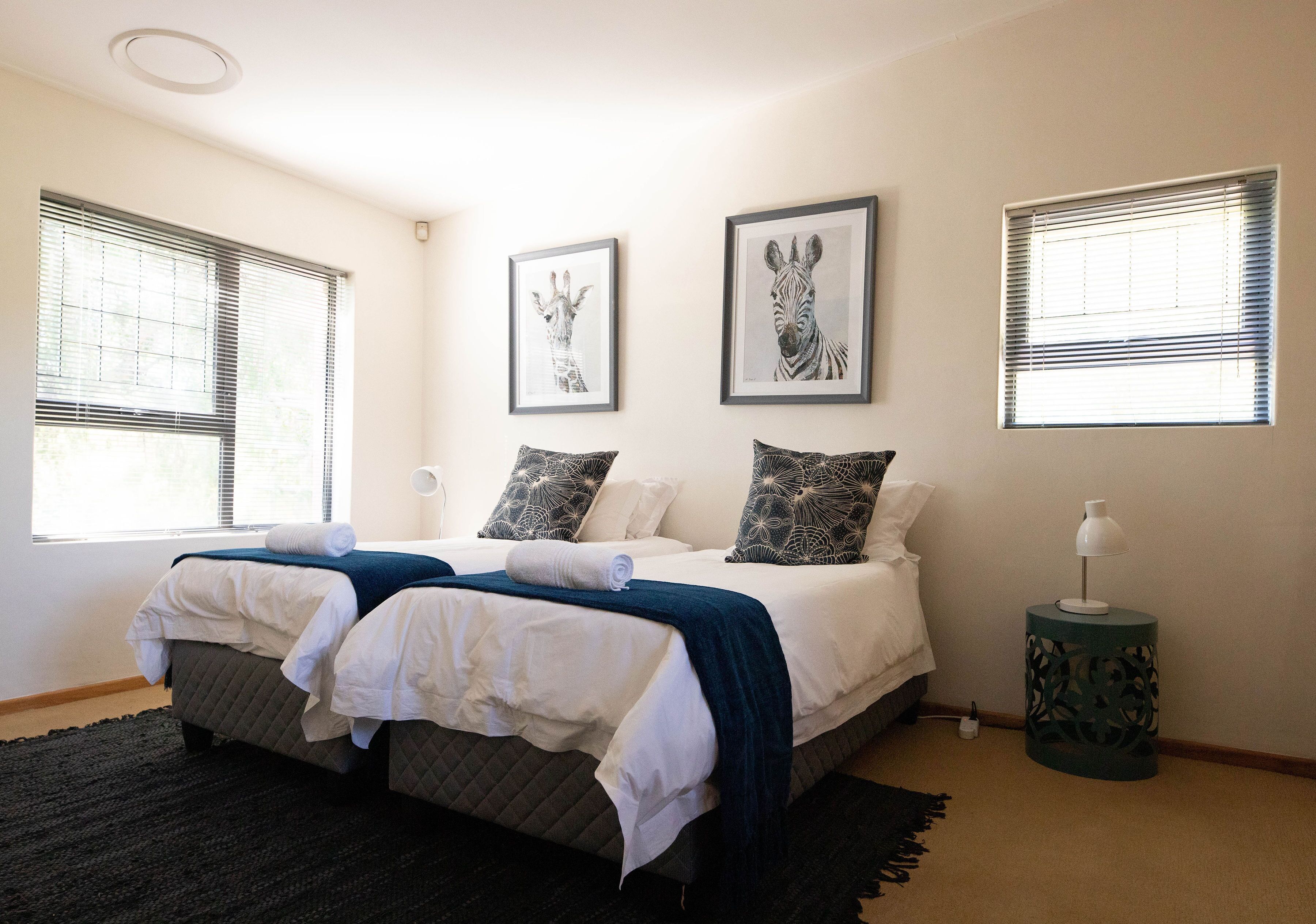 A double bed with white linens and a dark blue blanket, flanked by two framed animal prints, a small side table with a lamp, and a dark rug.