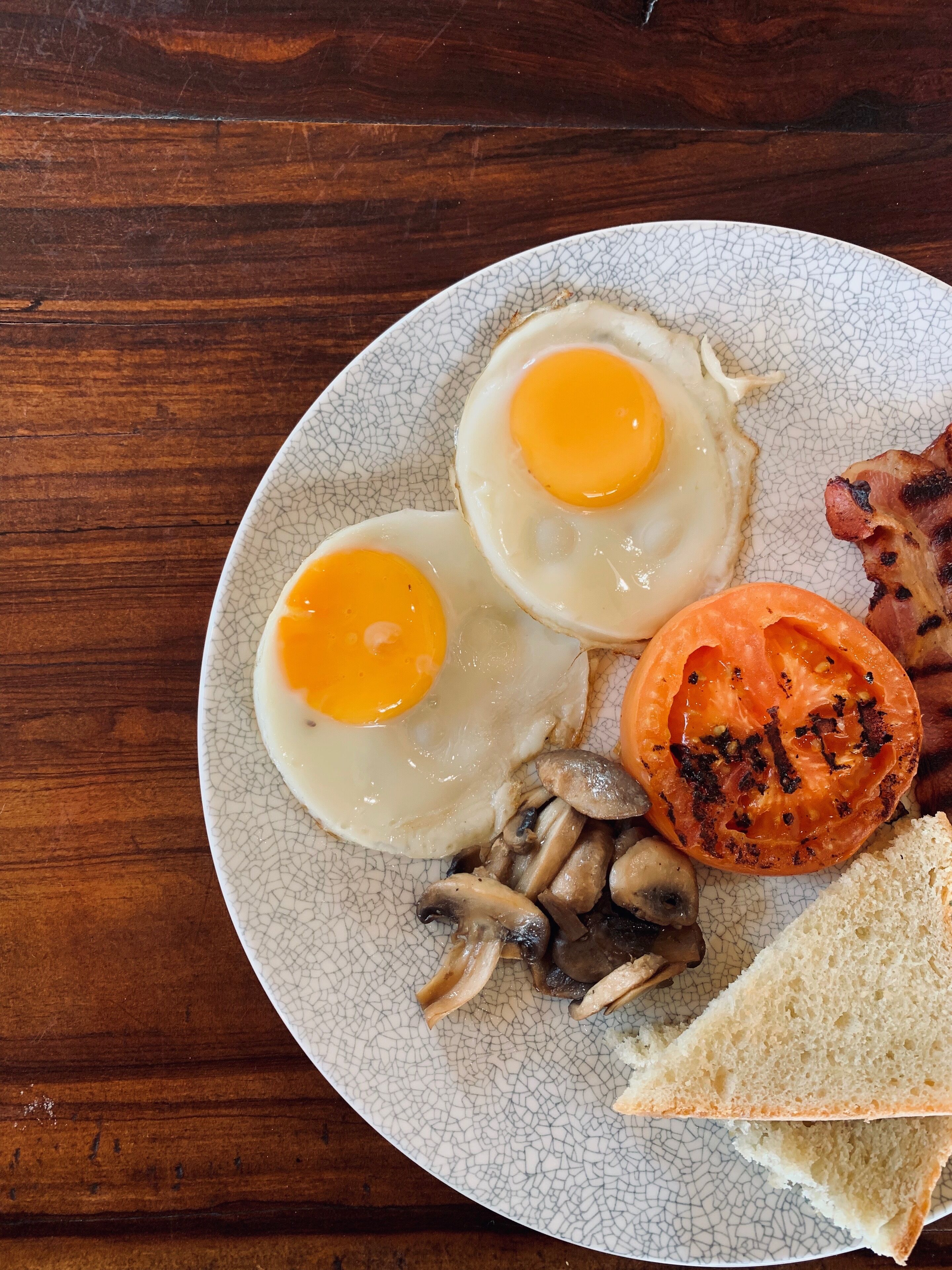 A plate with two sunny-side-up eggs, a grilled tomato, mushrooms, bacon, and a slice of toast.