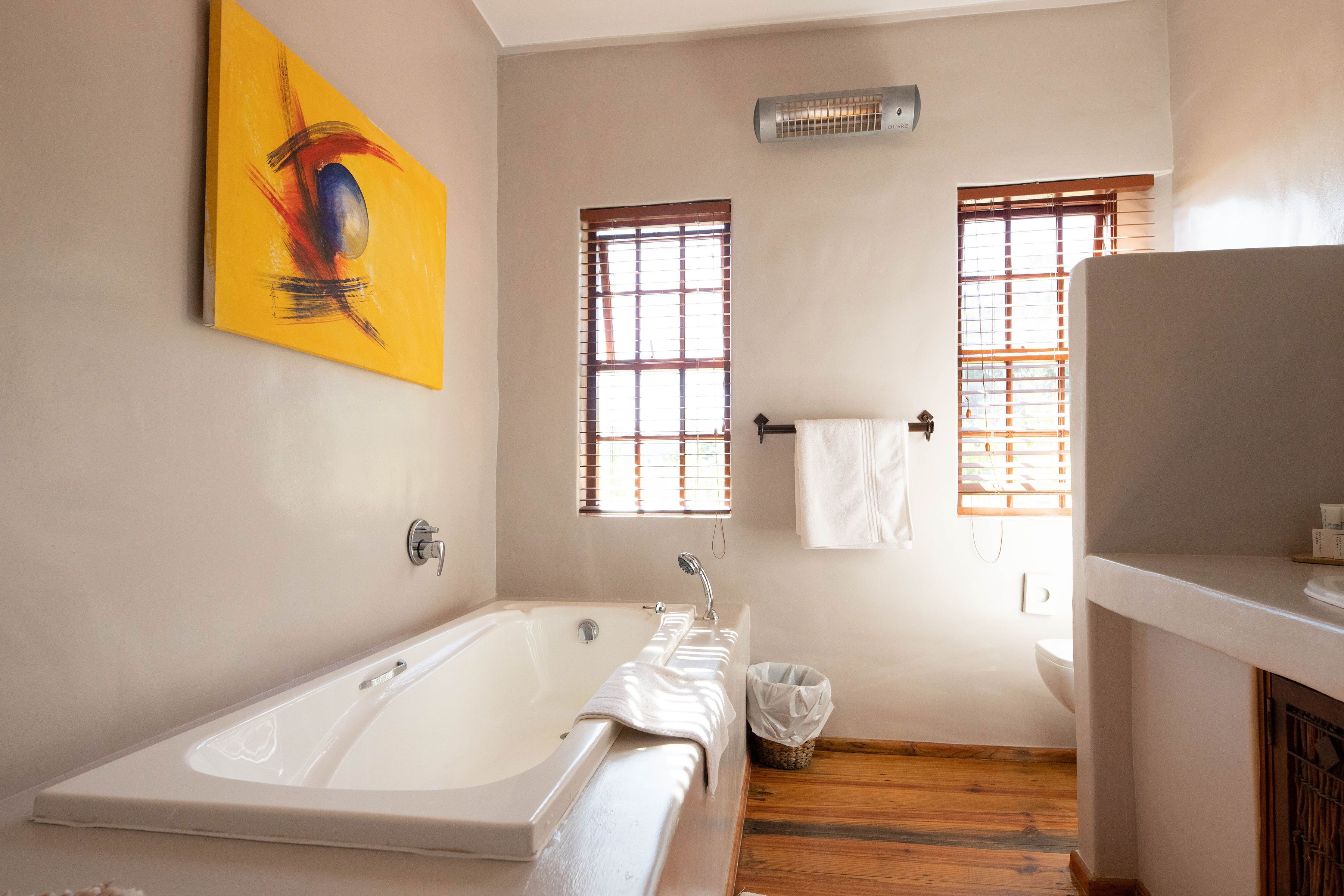 Un baño con bañera, dos ventanas con persianas, un perchero y un cubo de basura.