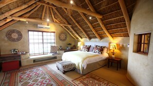 A bedroom with a sloped ceiling, a large bed, a window with blinds, a red bench, and a patterned rug. - Marianne Wine Estate (Stellenbosch)