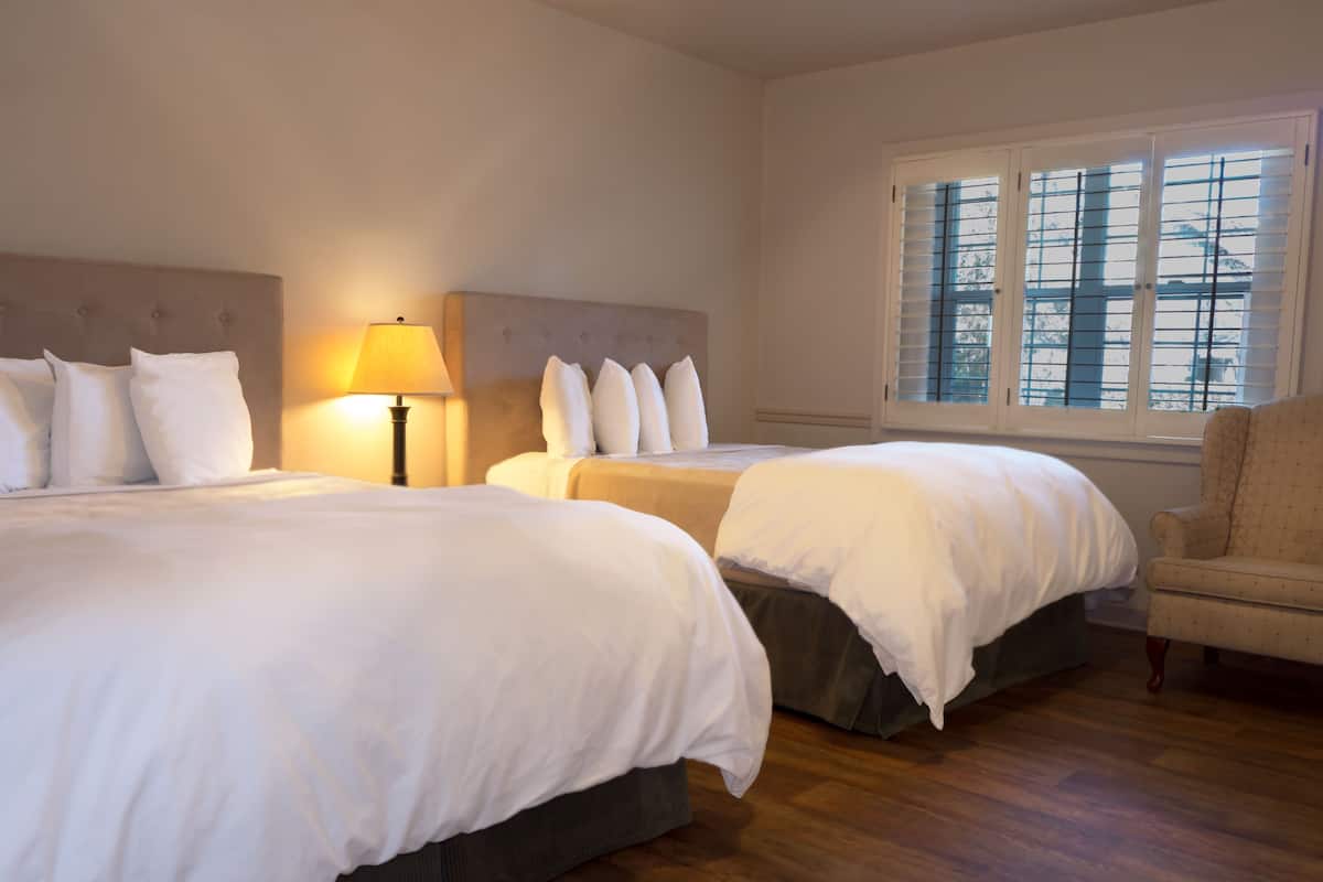 Ein Hotelzimmer mit zwei Betten, einem Nachttisch mit Lampe, einem Fenster mit Jalousien und einem Sessel.